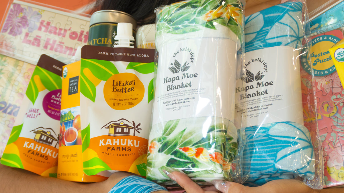A person holds multiple packaged Kaukuku products, including blankets and snack pouches, against a bright outdoor background. Visible products include a white Kapa Moe Blanket, a blue patterned blanket, a yellow Mac-Nut Crumble pouch, and colorful snack boxes.