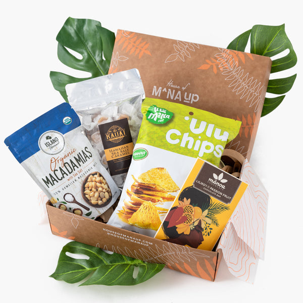 A gift set of Hawaiian snacks including packages of macadamia nuts, sea salt caramel candies, a chocolate bar, and 'Ulu chips, presented in a decorative box with tropical leaves.