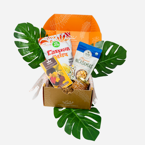 A curated gift box containing various snacks and treats, styled with large green monstera leaves on a light gray background.