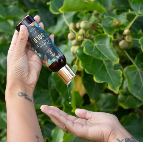 A hand holds a dark amber bottle of Maui Miracle Oil, tilting it to show a clear oil droplet dripping onto another hand, with green leaves and small green fruits in the background.