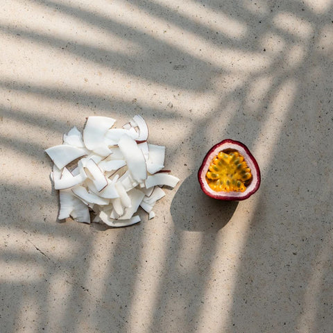 Coconut pieces and a halved passion fruit on a sandy surface with shadow patterns