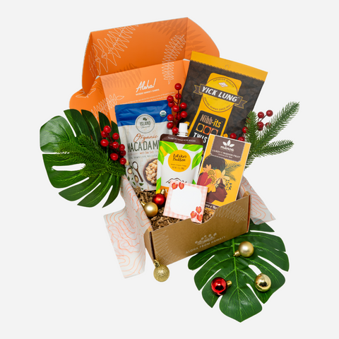 A curated gift box filled with assorted packaged snacks and treats, styled with green monstera leaves, red berries, pine branches, and gold ornaments on a light gray background.