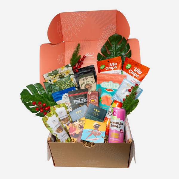 A curated gift box filled with assorted snack bags, packaged foods, a pink soda can, and green tropical leaves arranged inside an open kraft box on a light gray background.
