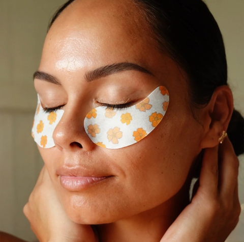 A close-up of a woman with closed eyes wearing white eye patches decorated with a yellow-orange floral pattern, with her hands gently touching her face.