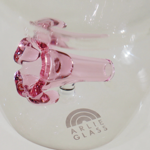 Pink glass puakenikeni on a white surface with 'Arlie Glass' branding.