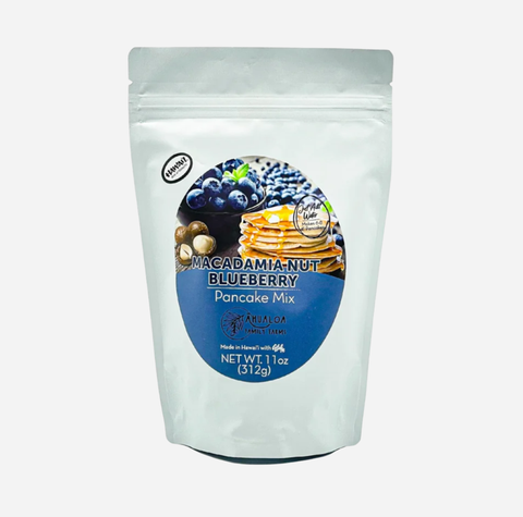 A stand-up pouch of Magadamia-Nut Blueberry Pancake Mix on a light gray background. The packaging is white with a large blue circular label featuring an image of blueberries, pancakes, and nuts.