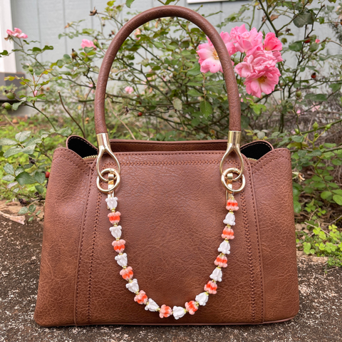Brown handbag with a decorative strap featuring pink and white beads, set against a natural background with flowers.