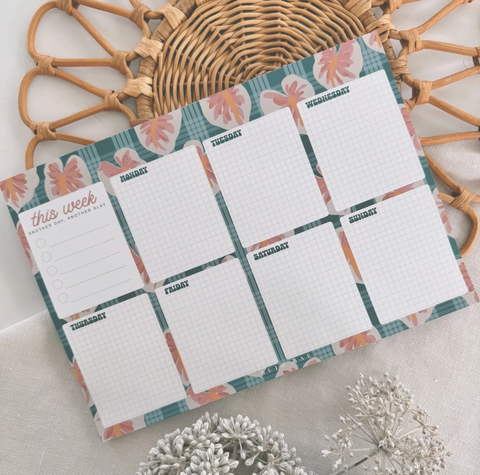 A weekly planner notepad with a teal plaid and floral border, laid flat on a light fabric surface with a wicker tray and dried flowers as props.