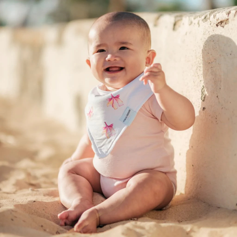 Baby sitting on sand wearing a pink outfit and floral bib.