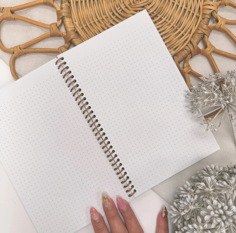 A white dotted grid planner or notebook with a silver spiral binding, shown open on a light fabric surface with decorative dried flowers and a woven tray around it. A hand with pink and gold manicured nails is visible at the bottom edge.