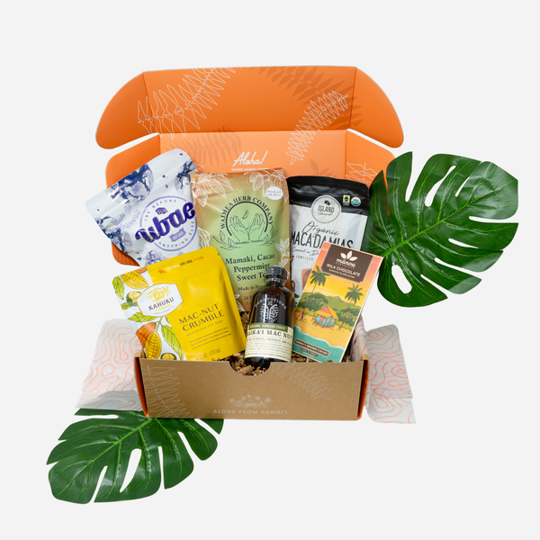 An open gift box filled with assorted packaged snacks and tea, styled with green monstera leaves on a light gray background.