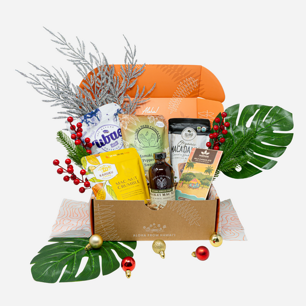 An open gift box filled with assorted packaged snacks and treats, styled with tropical leaves, silver pine branches, red berries, and small holiday ornaments on a light gray background.