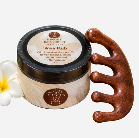 Hawaiian Rainforest Naturals 'Awa Rub facial massage scrub in a round wooden jar with a black lid, shown next to a carved wooden five-finger massage tool and a white plumeria flower on a white background.