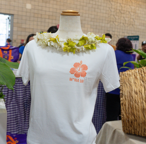 White short-sleeve T-shirt displayed on a mannequin torso at an indoor event, featuring a coral hibiscus flower graphic, with a white and yellow flower lei draped around the neck area.