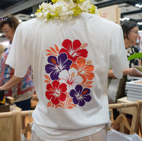 A white short-sleeve T-shirt shown from the back, featuring a large colorful hibiscus floral print in red, purple, and orange. The shirt is displayed on a mannequin or person in a busy indoor event or market setting, with a white flower lei draped over the shoulders.
