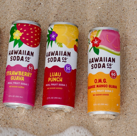 Three cans of Hawaiian Soda Co. real fruit soda arranged on sand: Strawberry Guava, Luau Punch, and O.