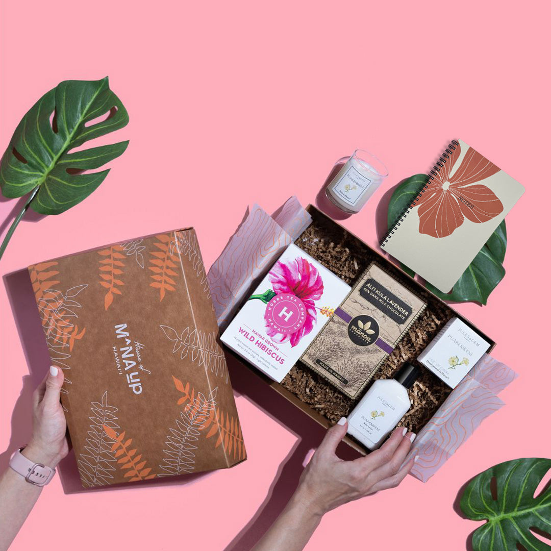 A gift set including a Bradley & Lily hibiscus large spiral notebook, Jules + Gem 2 oz soy candle and body lotion, and M\u0101noa Chocolate N\u0117nole, Hawai\u011i Island Bar 70%. Presented on a pink background with decorative leaves.