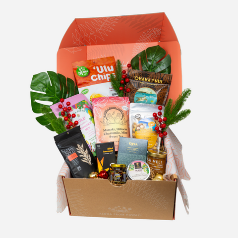 A curated gift hamper box filled with assorted packaged snacks, tea, and treats, decorated with green leaves and red berries, set against a plain light gray background.