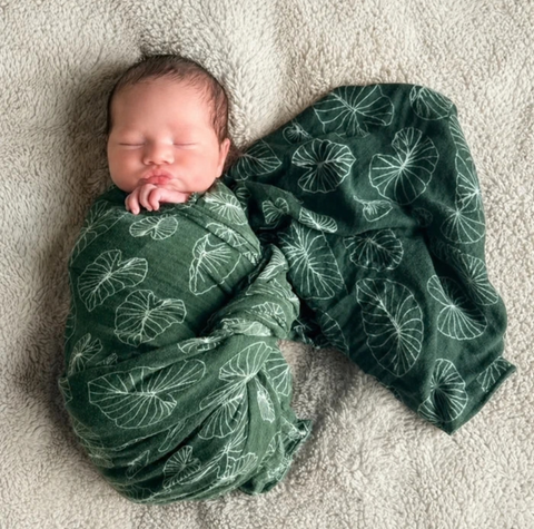 A sleeping newborn baby swaddled in a dark green muslin blanket with a white leaf pattern, lying on a textured beige blanket.