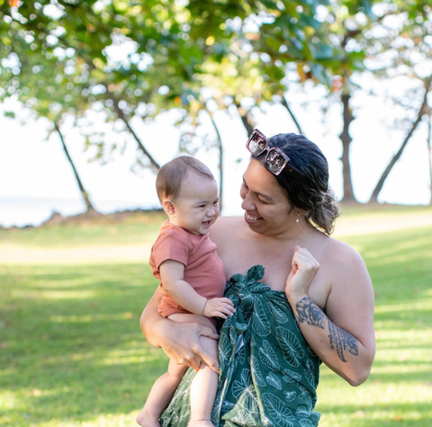 A woman outdoors holding a baby, wrapped in a dark green leaf-patterned sarong. The woman has sunglasses on her head and a tattoo on her arm, set against a background of green grass and trees.