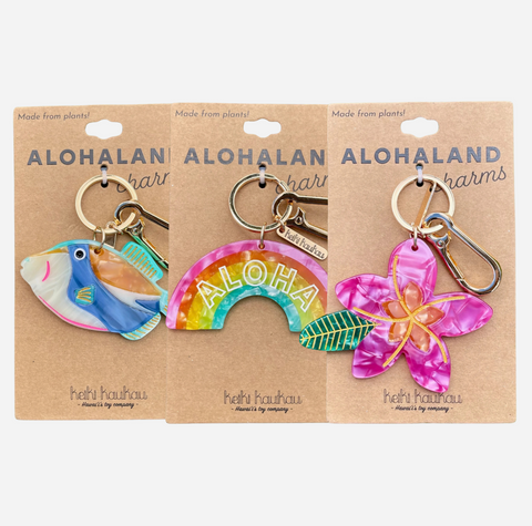 Three Alohaland charms on kraft paper backing cards. The set includes a blue tropical fish keychain, a rainbow arch with 'ALOHA' in the center, and a pink plumeria flower keychain.