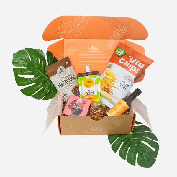 An open gift box filled with assorted packaged snacks and drinks, styled with large green monstera leaves on a light gray background. Visible items include a bag of macadamia nut, a bag of ulu Chips, a small bottle, and other snack packages.
