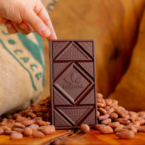 A hand holds a dark chocolate bar embossed with the brand name 'mahoia' against a warm background of cocoa beans and a burlap sack.