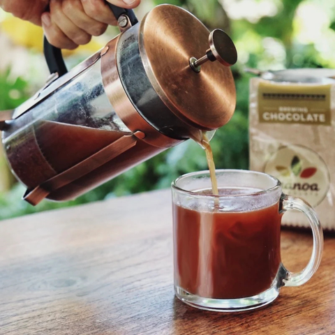 A hand pours freshly brewed coffee from a copper-and-stainless steel French press into a clear glass mug on a wooden table, with a bag of chocolate-flavored coffee beans blurred in the background.