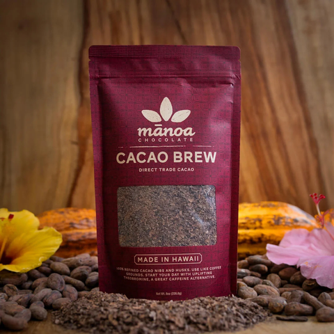 A stand-up pouch of Manoa Chocolate Cacao Brew direct trade cacao, set on a wooden surface surrounded by cacao beans, cocoa pods, and tropical flowers.