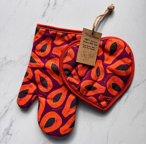 A two-piece quilted oven mitt set by Meli wraps, featuring a vibrant orange and purple papaya print. One mitt is laid flat showing the full pattern, and the other is folded over to display the quilted texture and red border.