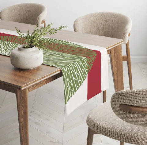 A rectangular table runner with a green and red geometric pattern and white border, styled on a wooden dining table with a small potted plant, surrounded by neutral upholstered chairs.