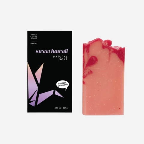 A bar of Sweet Hawaii Natural Soap in Tropical Plumeria, shown next to its black packaging box with purple geometric accents on a light gray background.