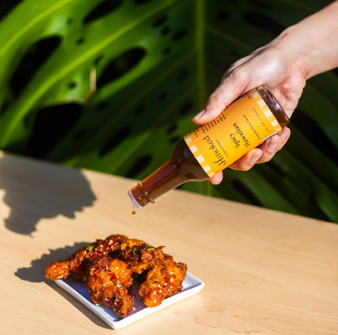 A hand holds a bottle of ponzu sauce and drizzles it over crispy fried chicken on a small white plate, set on a light wood table with a large green monstera leaf in the background.