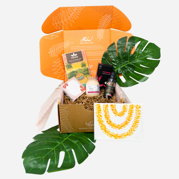 An open gift box from featuring tropical-themed items including a Hawaiian-themed candle, a small bottle, a card, and a floral print card, styled with green monstera leaves on a white background.