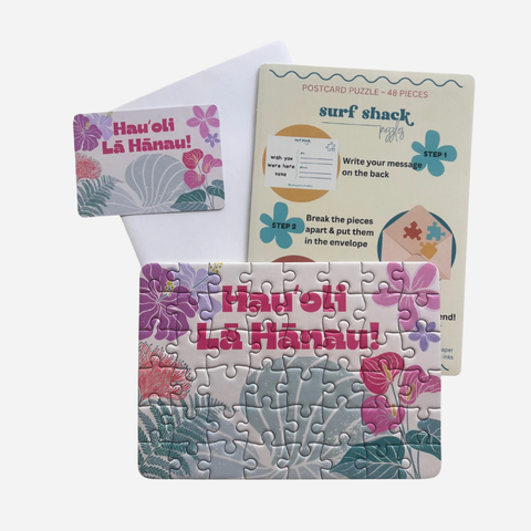 A postcard-style jigsaw puzzle set with floral tropical artwork and the text 'Hau'oli La Hánau!' shown both assembled and with the instruction card and envelope behind it on a light gray background.