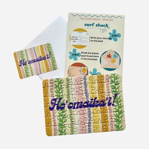 A postcard puzzle set featuring a completed jigsaw puzzle with the Hawaiian phrase 'Hōʻomāʻaila!' across a striped multicolor card design, shown alongside the instruction card and a matching envelope on a light gray background.