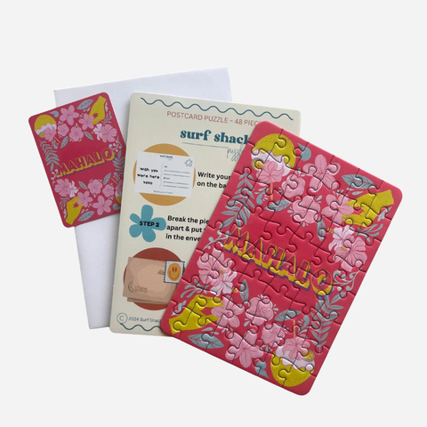 A pink postcard-sized jigsaw puzzle with floral and wave illustrations, shown alongside its instruction card and white envelope on a light gray background.