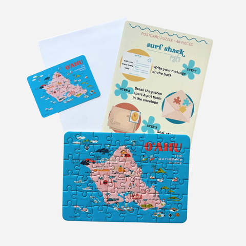 A postcard jigsaw puzzle set featuring a colorful illustrated map of Oahu, Hawaii, shown with a completed puzzle, an instruction card, and a white envelope on a light gray background.