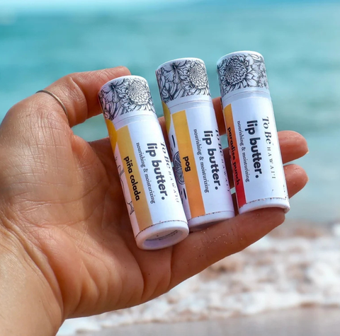Three lip butter tubes held in a person's hand with a beach and ocean background. The lip butters have white labels with yellow, orange, and red color blocks and black floral caps.