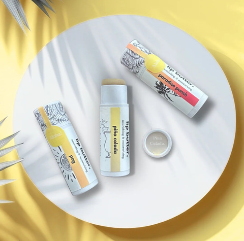 Three lip butter products from Pina Colada are arranged on a white circular tray with a yellow background. The products include a 'pina colada' lip butter stick, a 'paradise beach' lip butter stick, and a small round cap, all with white and yellow packaging and black illustrations.