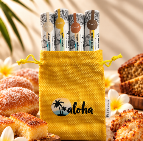A yellow drawstring pouch with the word 'aloha' printed on the front, holding four lip balm tubes. The pouch is surrounded by tropical food props, including sliced bread, sugar-dusted pastries, and white flowers, with a warm, sunlit background.