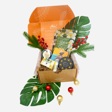 A curated gift box arrangement on a light gray background, featuring an open orange box with kraft filler, assorted packaged snacks and treats, green foliage, red berries, and hanging gold and red ornaments.