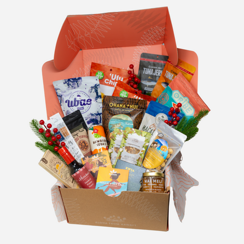 A curated gift box filled with assorted packaged snacks, teas, and treats, arranged in an open kraft cardboard box with a coral-orange inner lid.